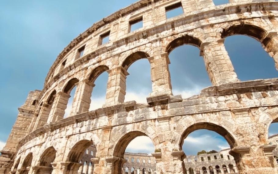 A close-up photo of the Pula Arena in Croatia. The arena is tan with tons of rounded open-air archways and through them you can see the clear sky. 